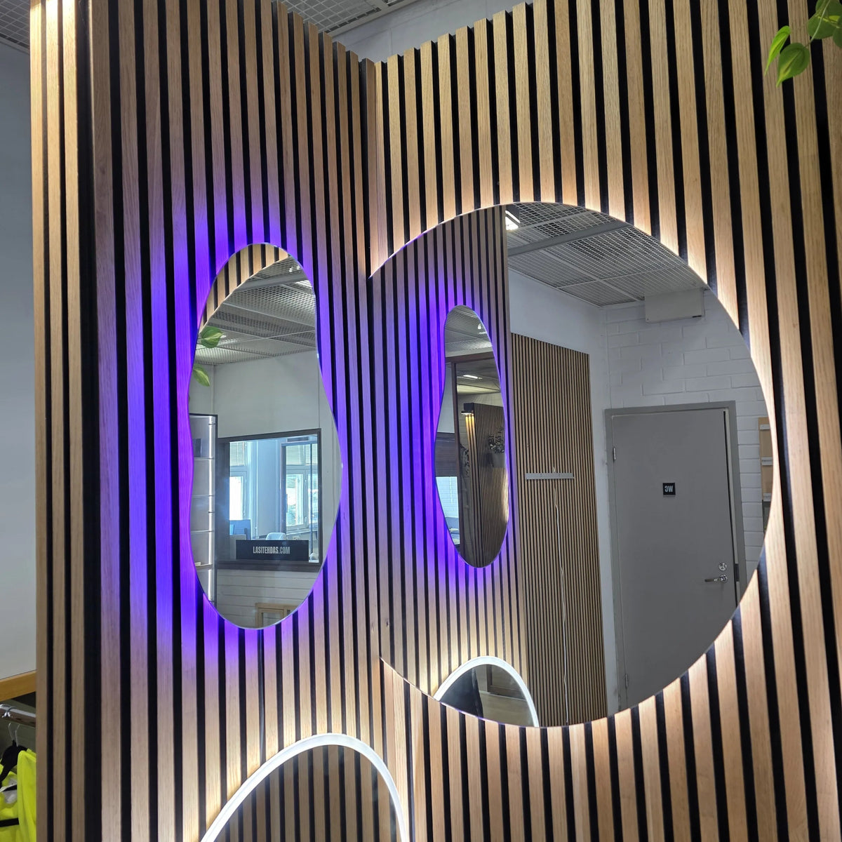 Puisella showroom-seinällä kaksi peiliä, toinen on epäsymmetrinen värivalovalaistu Hiekka-peili.